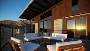A modern outdoor seating area with a sectional sofa, wooden lounge chairs, and a coffee table on a deck with large windows and a view of the hills. - Off-Grid, Modern Cliff House (Terlingua)