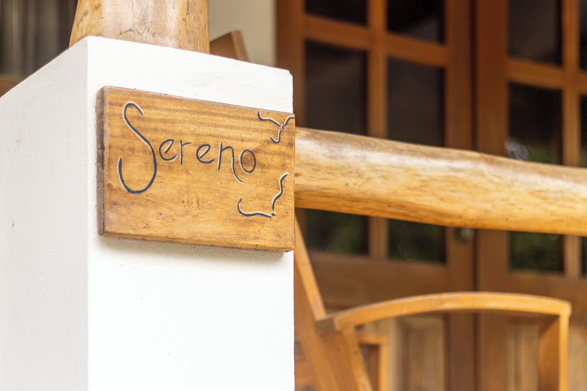 Ein Holzschild mit dem Schriftzug "Sereno" an einer weißen Wand, ein Holzstuhl und eine Holztür im Hintergrund.