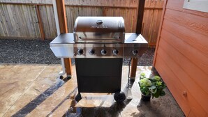 A stainless steel NEXGRILL with four burners, a digital display, and a side shelf, set up on a patio with a wooden fence and a house in the background. - Atargatis "the first mermaid"; an updated & spacious home, close to everything! (Brookings)