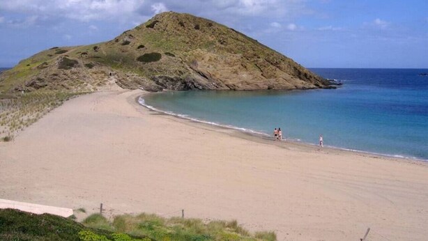 Une plage avec un rivage sablonneux, une petite colline et une eau bleue et calme.