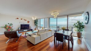 A modern living room with a large TV, a sofa, a dining table with chairs, and a view of the ocean through large windows.