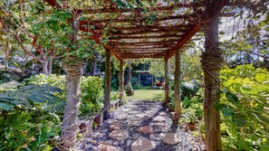 A garden with a wooden pergola structure, surrounded by lush greenery and a stone pathway.