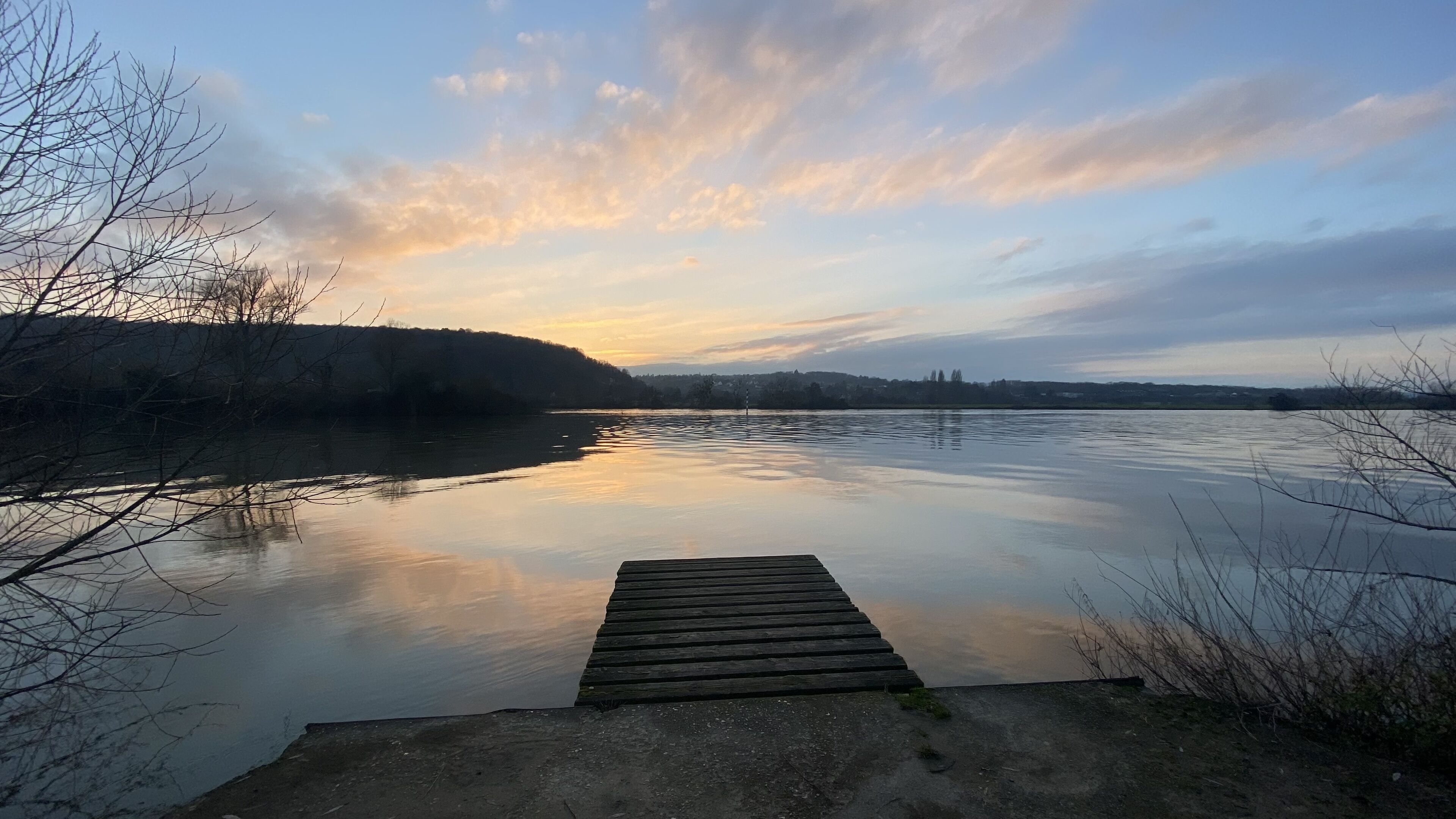 En træbro strækker sig ud i stille vand under en klar himmel.
