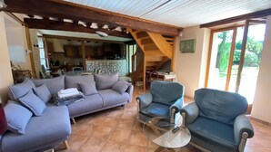 A living room with a sofa, two armchairs, and a glass coffee table. There is a stone wall and wooden beams in the ceiling. A kitchen is visible in the background. - The Nebula (notre dame de l'isle)