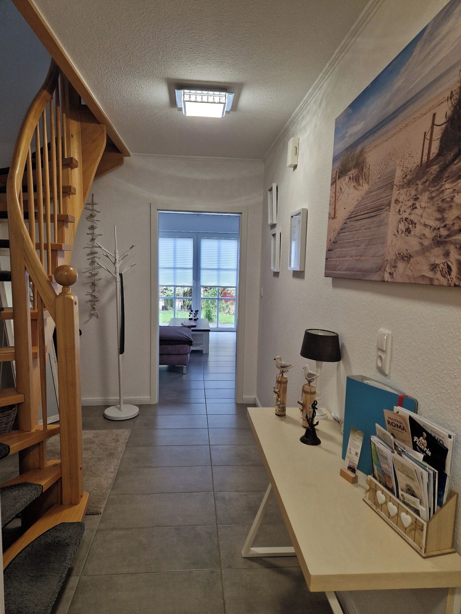 A home interior with a staircase, a desk with a lamp, a painting, and a view through a door to an outdoor area.