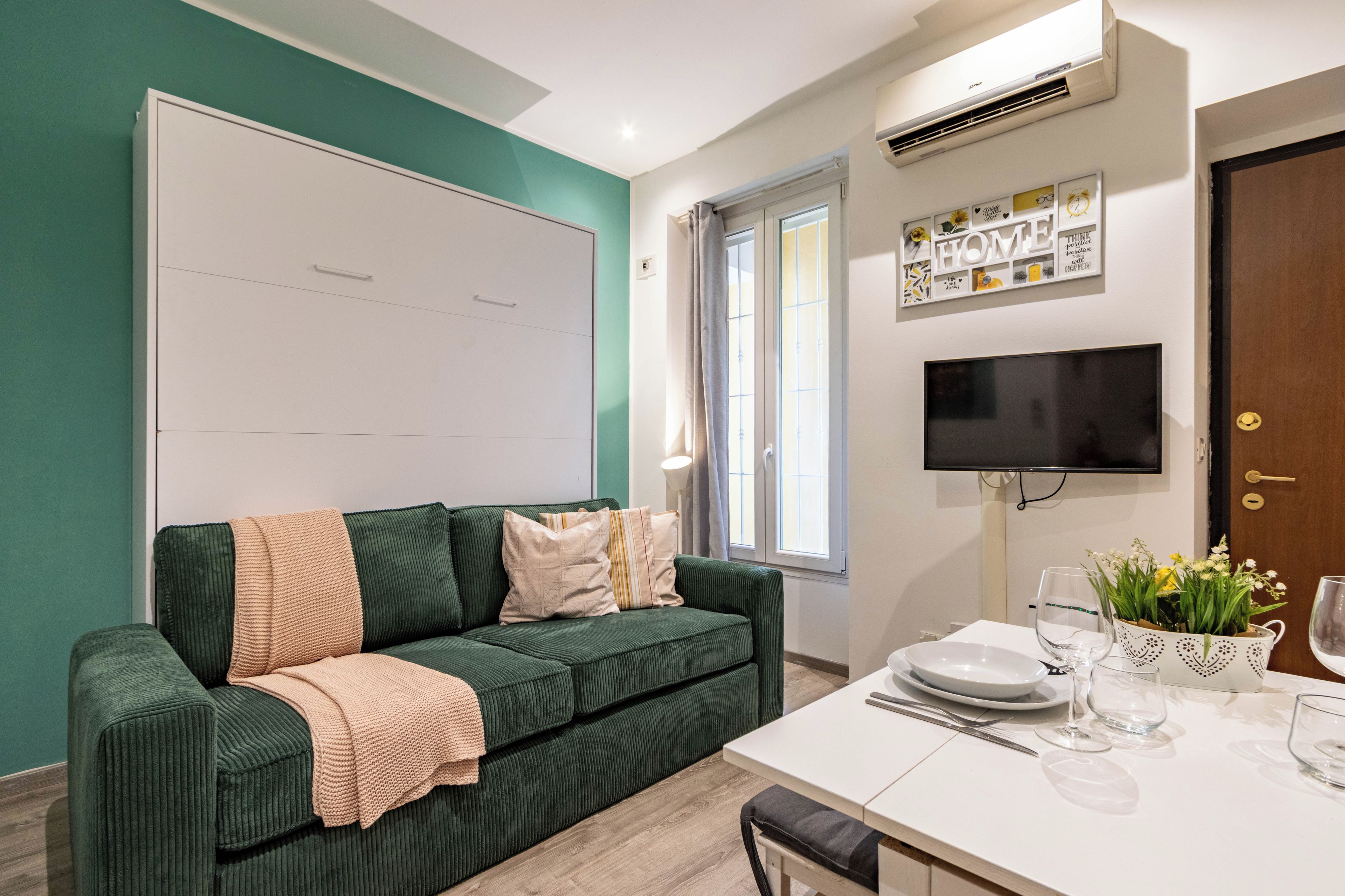 Un soggiorno moderno con un divano verde, un tavolo bianco apparecchiato per la cena, una TV a schermo piatto montata a parete e una finestra con tende.
