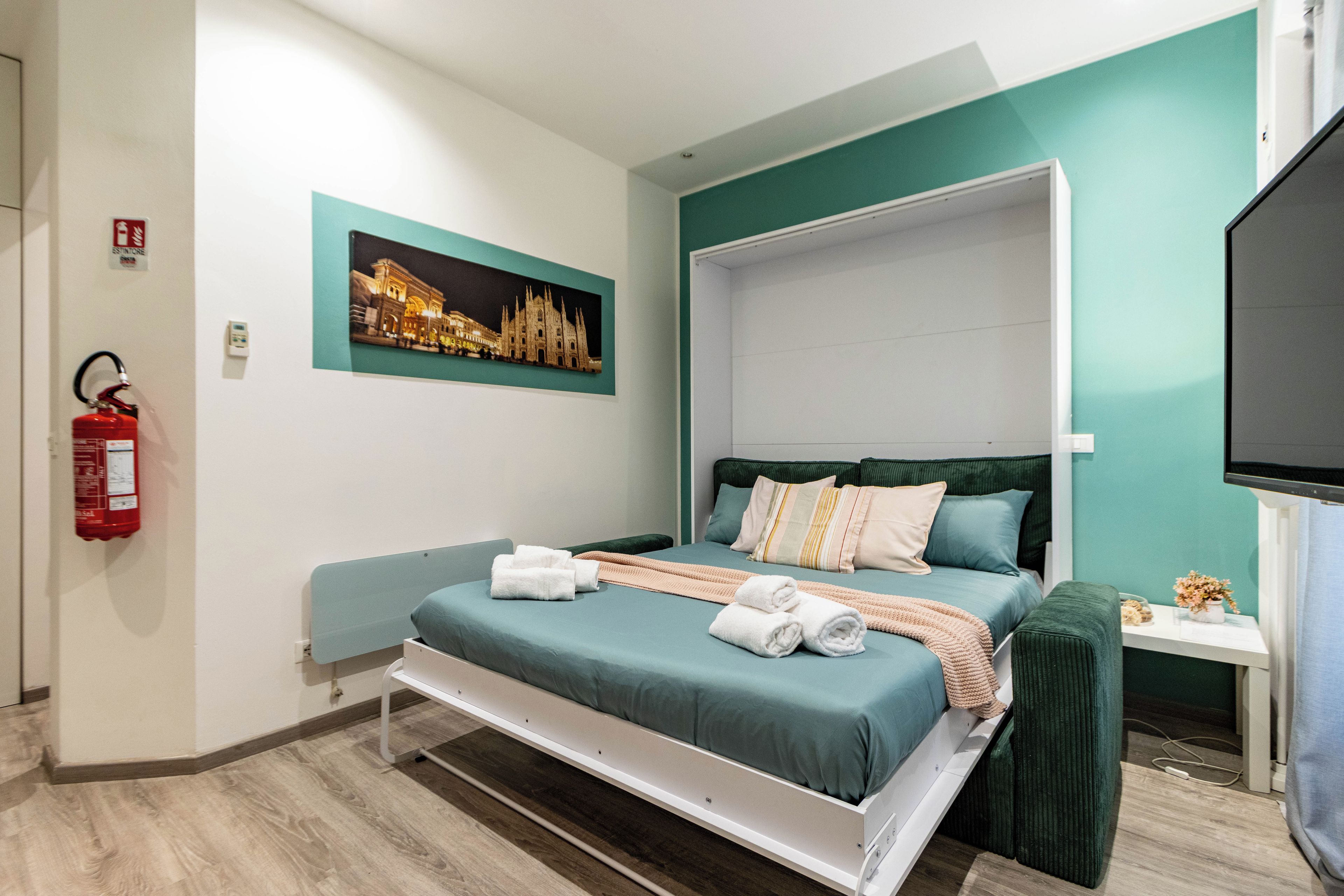 Una camera d'albergo con un letto, una TV a schermo piatto, un quadro raffigurante un edificio e un estintore.