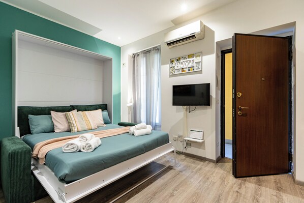 A hotel room with a bed, a television, a door, and a wall-mounted air conditioning unit.