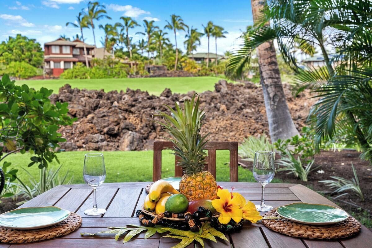 A wooden table set with fruits, a pineapple, a hibiscus flower, and two wine glasses, overlooking a volcanic landscape and a house.