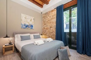Un dormitorio con cama, mesita de noche, cuadro en la pared, pared de piedra y vistas a través de una ventana con cortinas.