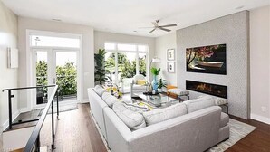 A modern living room with a grey sofa, a glass coffee table, and a fireplace. There is a balcony with a view of greenery. - STUNNING LUXURY BEACH HOUSE (Cleveland)