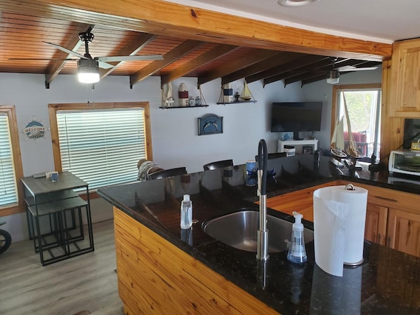 A modern kitchen with a black countertop, stainless steel sink, and wooden cabinets. There is a bar area with stools and a welcome sign.