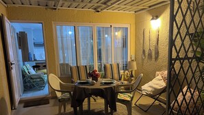 Comedor con una mesa dispuesta para cuatro personas, rodeada de sillas. Se observa una ventana con cortinas, una puerta que conduce a otra habitación y una lámpara fijada a la pared.