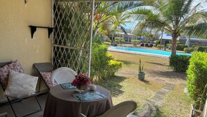 Un patio exterior con una mesa dispuesta para dos personas, rodeado de sillas, una piscina al fondo y vistas de un edificio con techo azul.