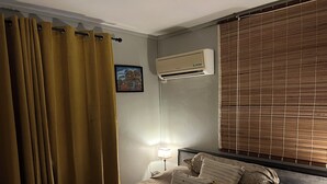 Un dormitorio con cama, cortinas, aire acondicionado, una ventana con persianas y una lámpara de noche.