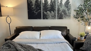 Un lit avec une literie à rayures, un plaid foncé et une tête de lit. Trois cadres photos représentant des scènes de forêt sont accrochés au mur. On aperçoit une lampe de sol et une petite table sur laquelle est posée une plante.