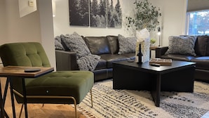Un salon moderne avec un fauteuil vert, un canapé en cuir noir et une table basse. Deux tableaux encadrés ornent le mur, ainsi qu’une plante en pot et une fenêtre avec des stores.