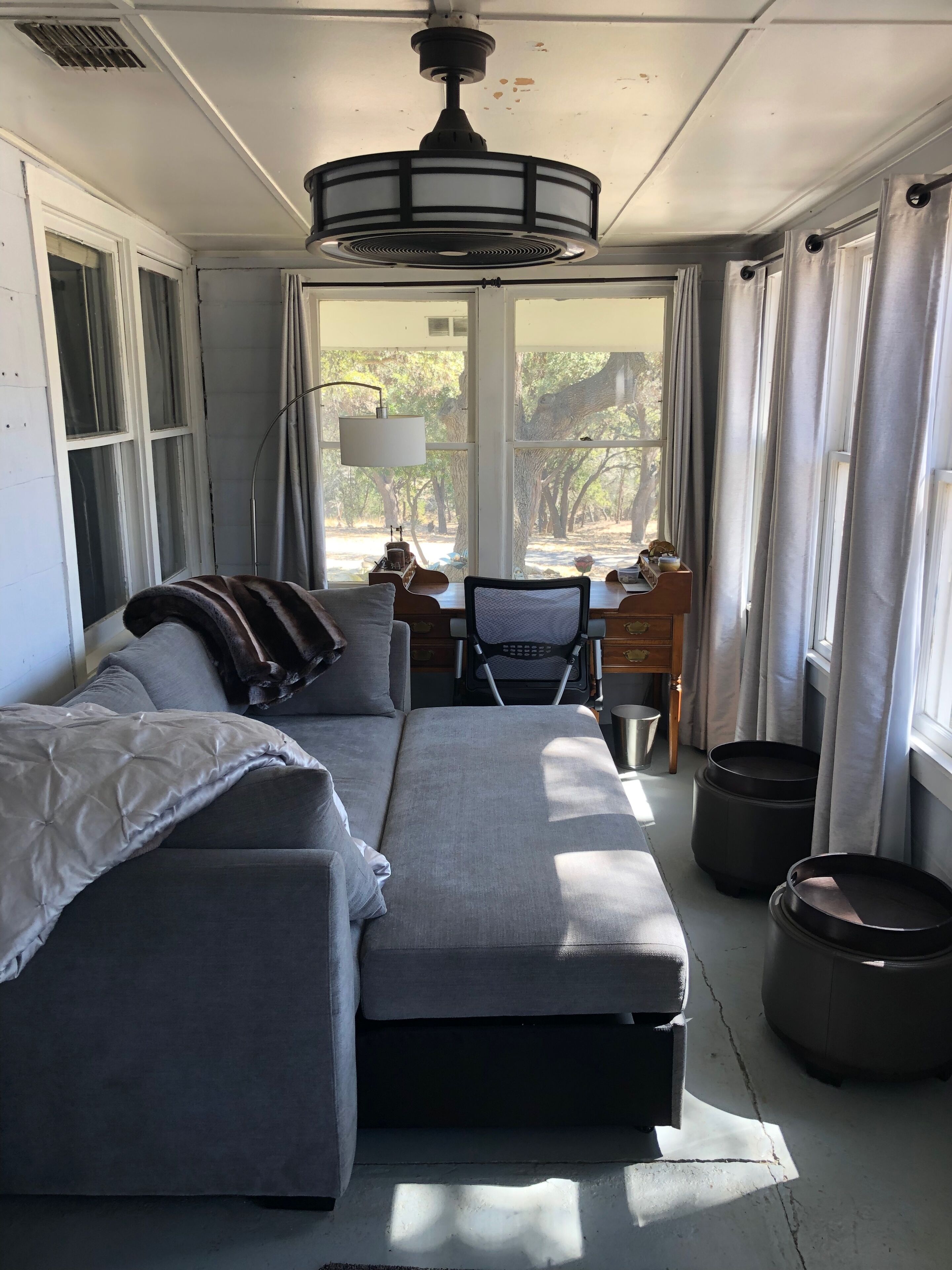A modern living room with a grey sectional sofa, a small table, and a chair. There is a ceiling fan, a window with curtains, and a view of trees outside.