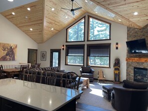 A spacious living area with a large wooden ceiling, a stone fireplace, and a large window offering a view of trees.