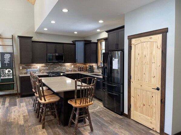 A modern kitchen with dark cabinets, a stainless steel refrigerator, a large island with stools, and a wooden door.