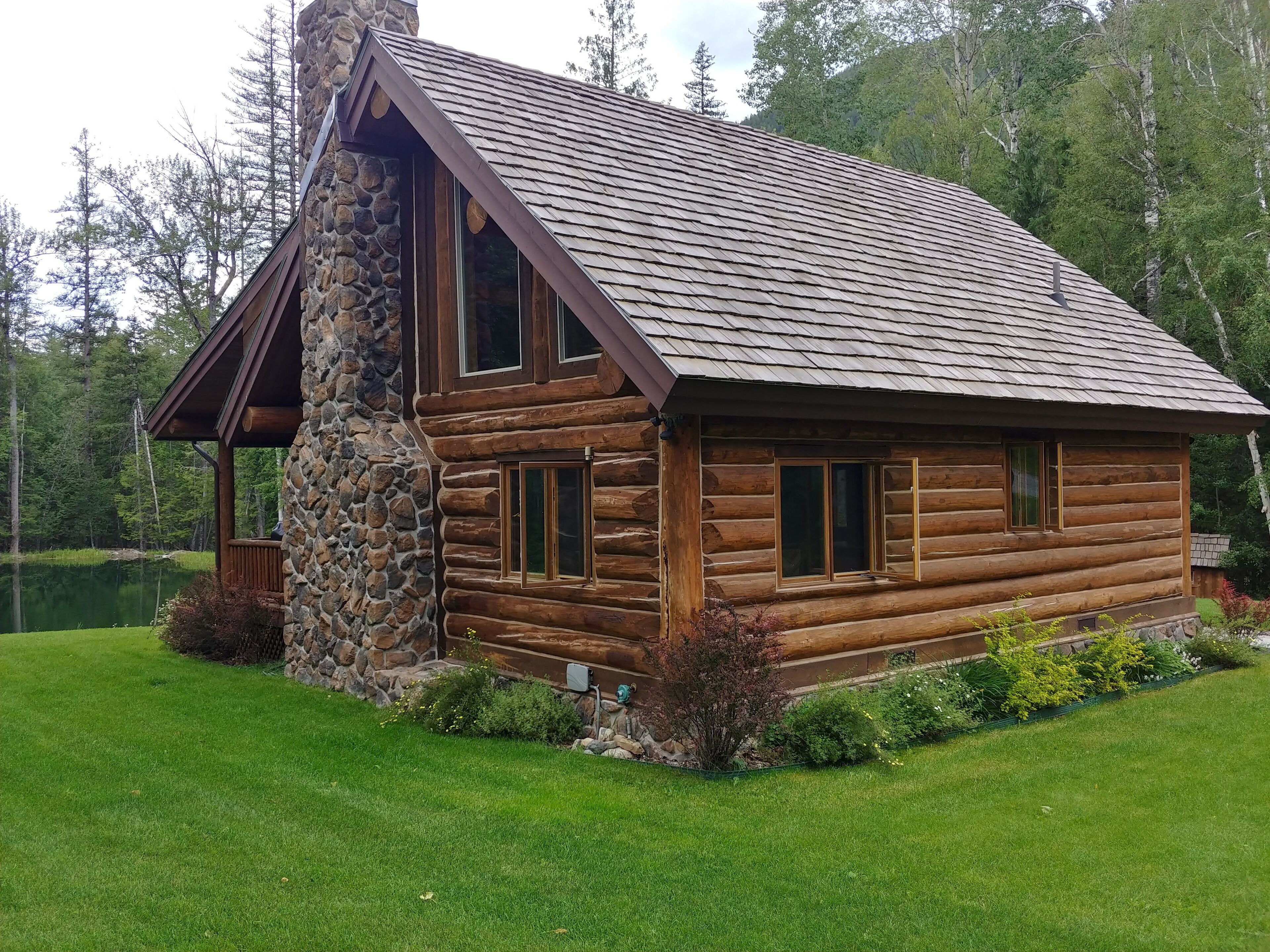 A log cabin with a stone chimney, large windows, and a view of a lake.