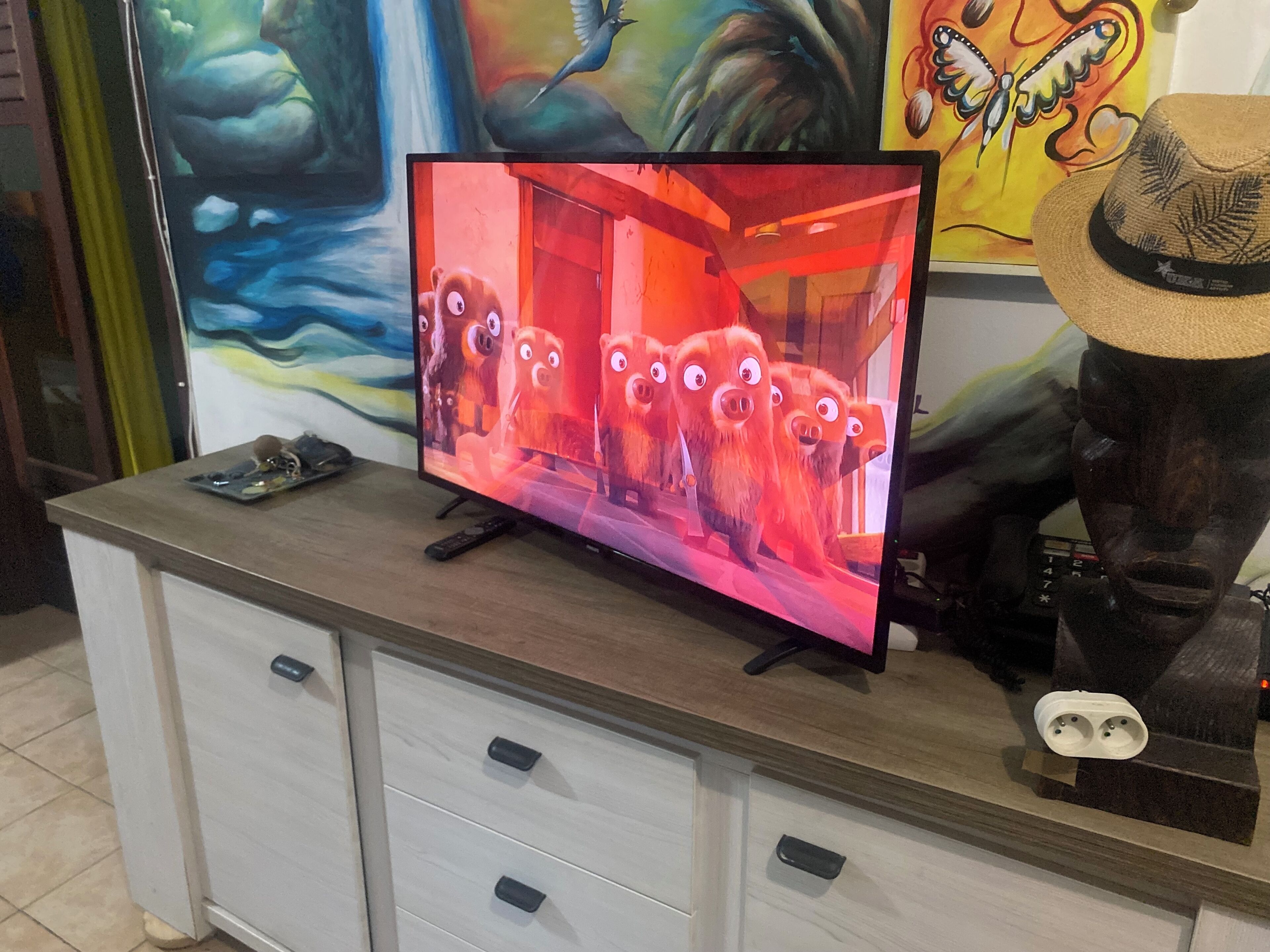 A television on a wooden cabinet with a remote control, a decorative hat, and a wall with colorful artwork.
