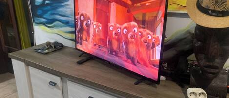 A television on a wooden cabinet with a remote control, a decorative hat, and a wall with colorful artwork.