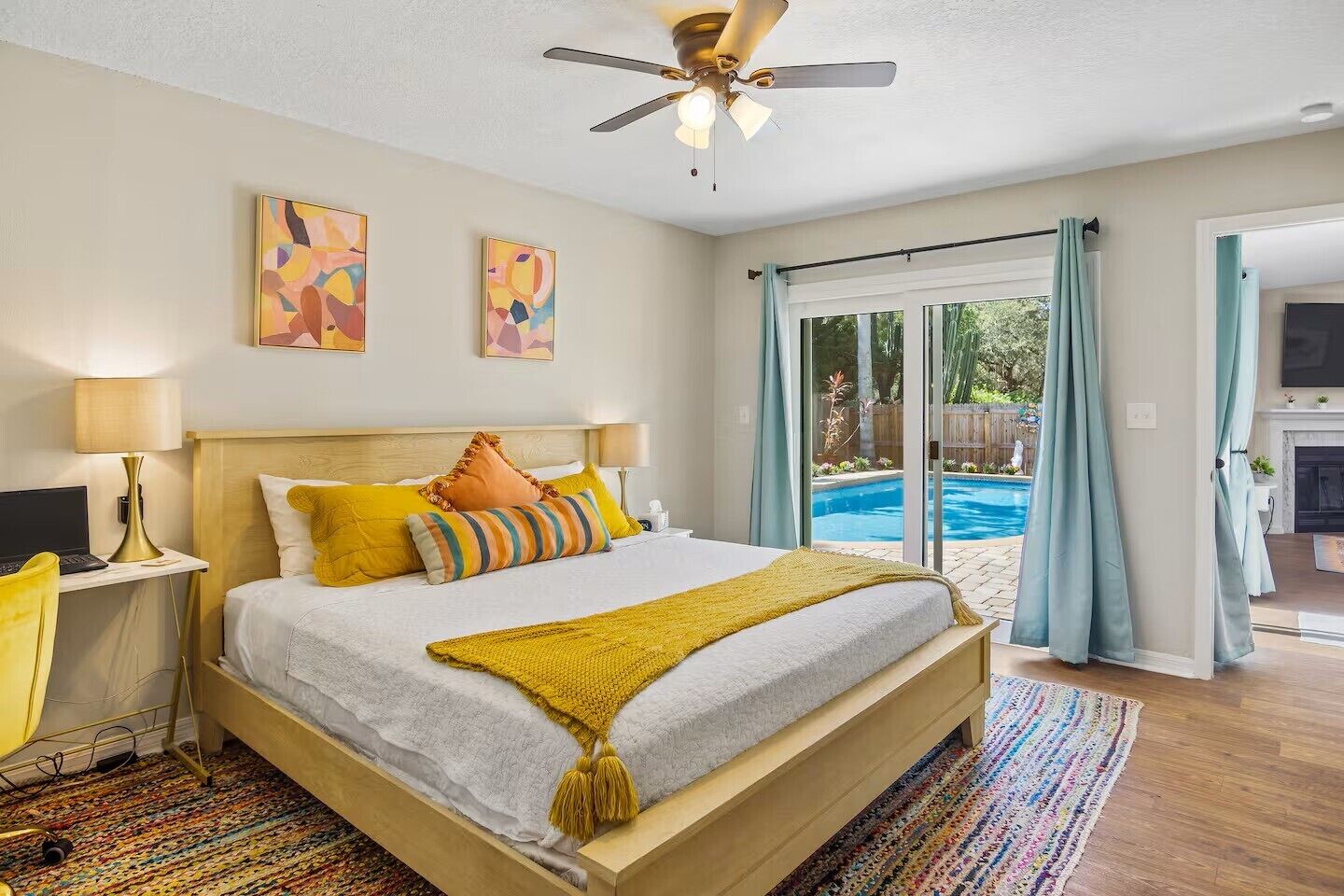 Un dormitorio con cama, escritorio con lámpara, ventilador de techo y vista a una piscina al aire libre.