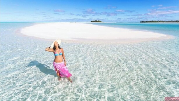Una donna in bikini e gonna rosa in piedi su una spiaggia di sabbia bianca con acqua turchese trasparente.