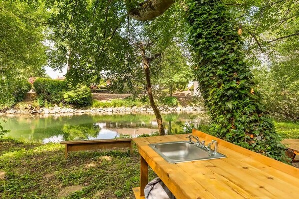 Taula de picnic de fusta amb una pica, envoltada de vegetació exuberant i un estany tranquil.