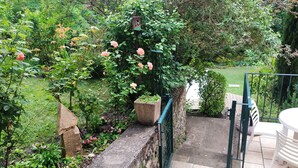 A garden with a stone wall, a wooden birdhouse, and a metal gate leading to a patio area with a white table and chairs. - 2* gîte on the ground floor of an old pottery in the center of Dieulefit (Dieulefit)