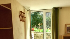 A room with a wooden chair, a tiled floor, a door leading outside, and a small table with a picture frame. - 2* gîte on the ground floor of an old pottery in the center of Dieulefit (Dieulefit)