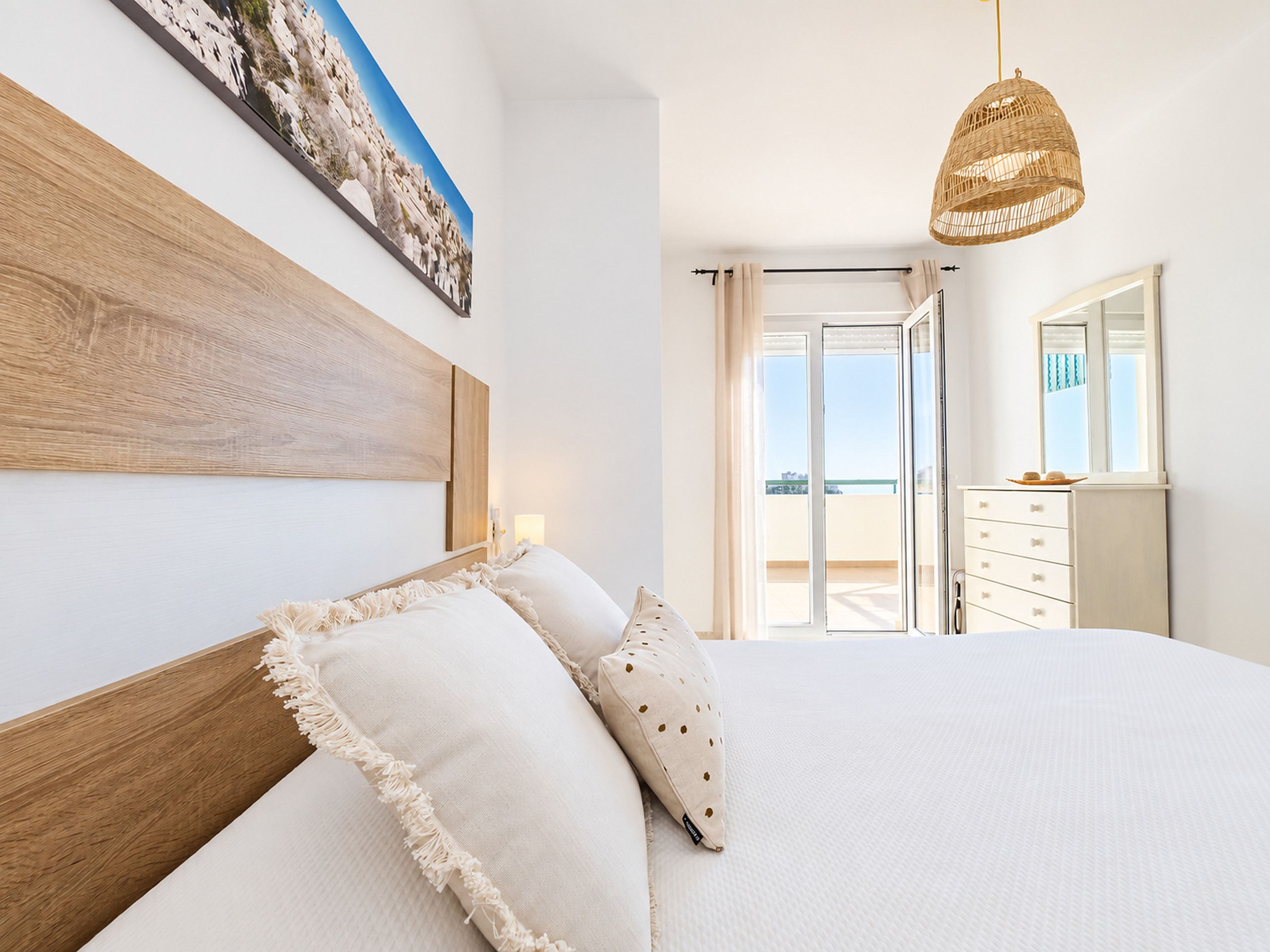 A bedroom with a large bed, a wooden headboard, a hanging rattan light fixture, and a view of the outdoors through a sliding door.