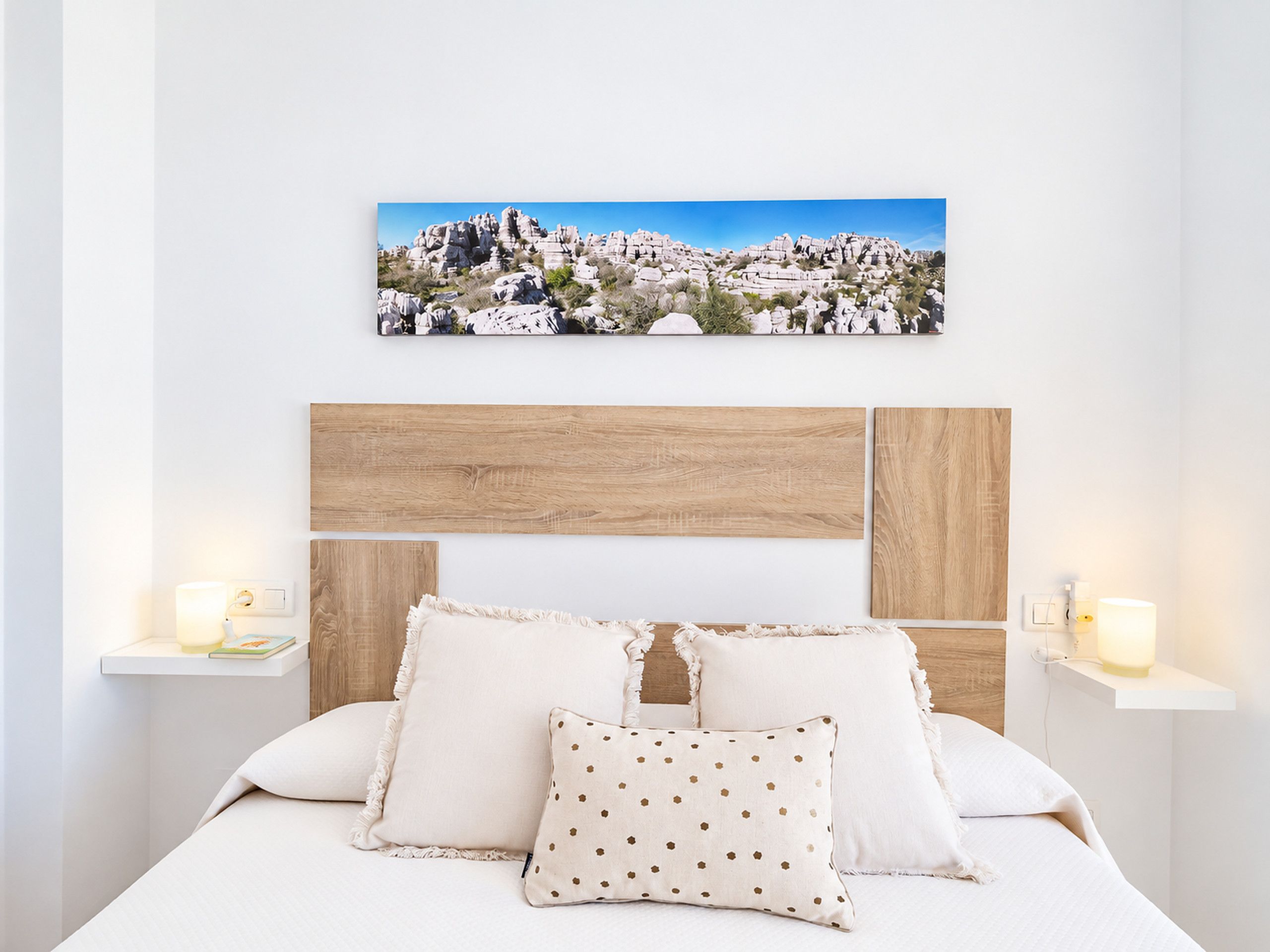 A bedroom with a bed, pillows, a wooden headboard, bedside tables, and a wall-mounted picture.