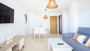 A modern living room with a grey sofa, white coffee table, and a TV mounted on the wall. There is a dining area with a white table and chairs, and a wicker pendant light above. - Cubo’s La Terraza del Mar / free wifi / sea views (Algarrobo)