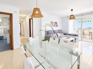 A modern living room with a glass dining table, grey sofa, and wicker pendant lights.