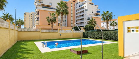 A swimming pool with a diving board, surrounded by a well-maintained lawn and palm trees, with a multi-story building in the background.