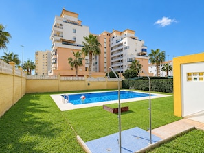 A swimming pool with a diving board, surrounded by a well-maintained lawn and palm trees, with a multi-story building in the background.