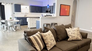 A modern living room with a grey sofa, two bar stools, and a dining table with chairs.