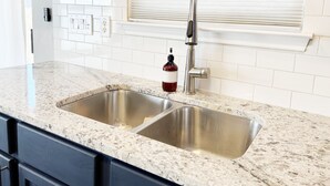 A kitchen with a double stainless steel sink, a granite countertop, a faucet, and a bottle of red liquid.