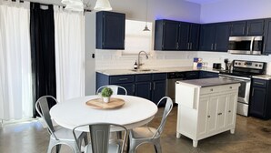 A modern kitchen with dark blue cabinets, a white countertop, and stainless steel appliances. There is a round dining table with metal chairs and a small potted plant.