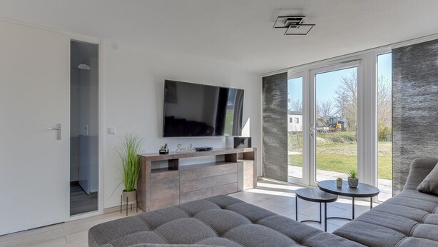 Un soggiorno moderno con un ampio divano grigio, una televisione a schermo piatto montata su un mobile in legno e una porta vetrata scorrevole che dà su un balcone.