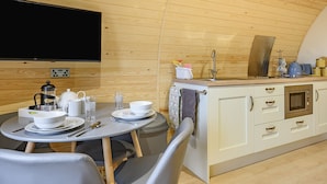A cozy, compact kitchen with wooden walls, a dining area with a table set for two, and a flat-screen TV mounted on the wall. - Unwind in a relaxing a fun accommodation in a beautiful rural setting. (East Haddon)