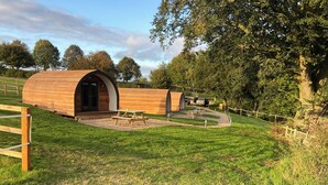 A series of wooden, semi-circular structures with picnic tables in a grassy area, surrounded by trees and a fence. - Unwind in a relaxing a fun accommodation in a beautiful rural setting. (East Haddon)