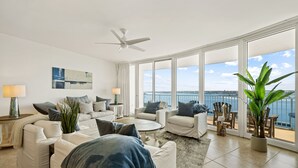 A spacious living room with a large window overlooking the sea, furnished with white sofas and a ceiling fan. - Newly Renovated Condo with Bay Views! Complimentary Boat Slip! (Orange Beach)