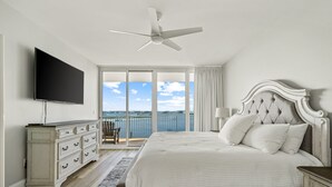 A bedroom with a bed, a flat-screen TV, a ceiling fan, a dresser, a nightstand, and a view of the sea through large windows. - Newly Renovated Condo with Bay Views! Complimentary Boat Slip! (Orange Beach)