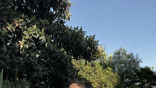 A person in a white robe holding a cup, standing in a garden with lush greenery and a clear sky.