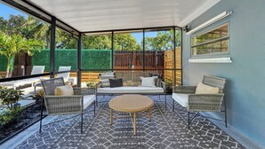Un porche cubierto con muebles de mimbre, una mesa redonda y vistas a un seto verde y una valla de madera.