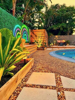 A pool area with a wooden deck, pebbles, and a neon rainbow light decoration. - Stylish Pet Friendly Home with Private Pool Oasis (Fort Lauderdale)