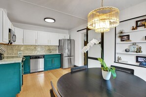 A modern kitchen with white cabinets, a teal island, stainless steel appliances, and a dining area with a dark table and chairs.